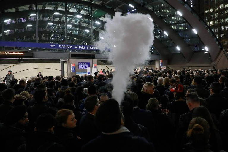 A imagem mostra uma grande multidão de pessoas na estação Canary Wharf， com um denso vapor saindo de uma fonte. O ambiente é urbano， com paredes de vidro e iluminação artificial. Ao fundo， há um painel informativo e a entrada da estação é visível. A maioria das pessoas está de pé， algumas olhando para o painel， enquanto outras conversam.