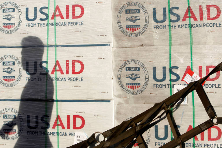 A imagem mostra a sombra de uma pessoa caminhando em frente a pilhas de caixas empilhadas， que têm a marca da USAID visível. As caixas estão organizadas e amarradas com fitas verdes， e a logo da USAID é destacada em vermelho e azul.