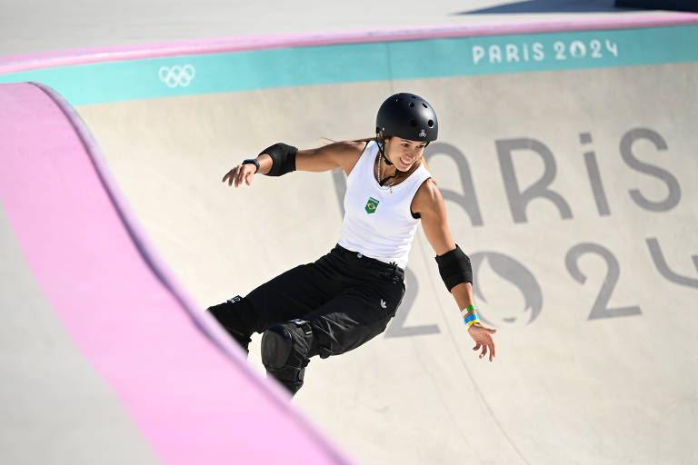 Uma skatista está realizando uma manobra em uma pista de skate. Ela usa um capacete preto e roupas esportivas， incluindo uma camiseta branca e calças pretas. A pista tem bordas rosa e o logotipo dos Jogos Olímpicos está visível no canto superior esquerdo. Ao fundo， está escrito 039;PARIS 2024039;.
