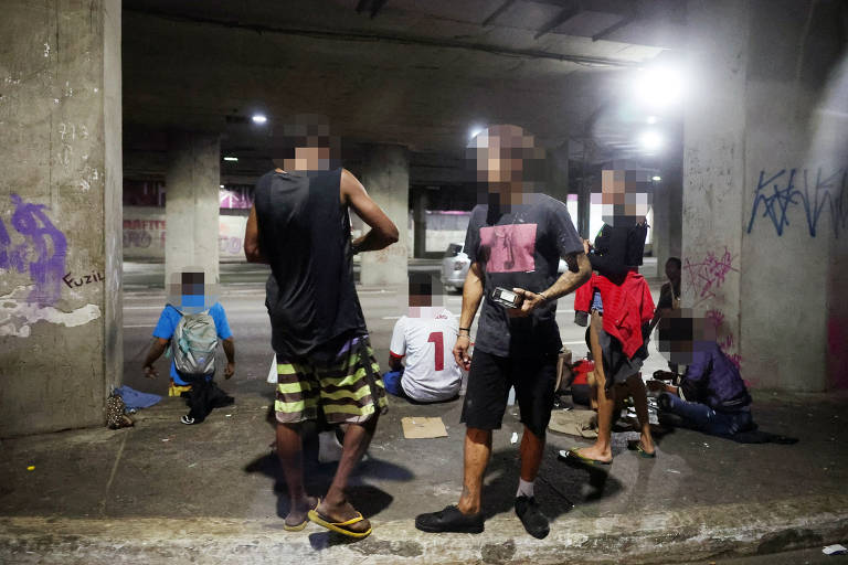 A imagem mostra um grupo de pessoas em uma área coberta. Algumas pessoas estão sentadas no chão, enquanto outras estão em pé. O ambiente é mal iluminado, com luzes artificiais visíveis. As paredes têm grafites