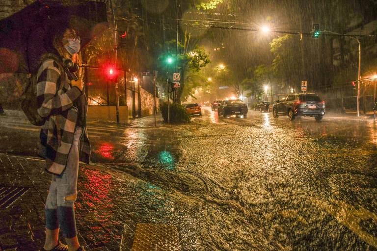 Uma mulher está parada em um cruzamento durante uma forte chuva. Ela usa uma máscara e segura um guarda-chuva. O cenário é iluminado por luzes de semáforo vermelhas e verdes， e há carros passando na rua alagada. A chuva é intensa， criando um efeito de água acumulada no chão.