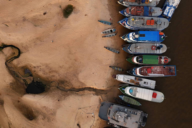 A imagem mostra uma vista aérea de uma área de praia com areia e água. À direita， há uma fileira de barcos de diferentes tamanhos e cores， ancorados na margem. A areia é de tonalidade clara e há algumas manchas escuras na água. À esquerda， um pequeno curso de água serpenteia pela areia.