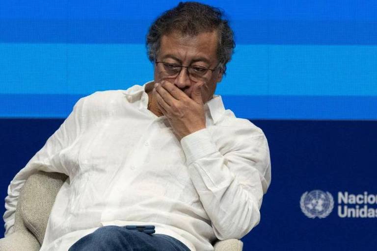 Um homem sentado em um palco， com uma expressão pensativa， cobrindo parcialmente o rosto com a mão. Ele usa uma camisa branca e jeans. O fundo é composto por faixas horizontais em tons de azul. No canto inferior direito， há o logotipo das Nações Unidas.
