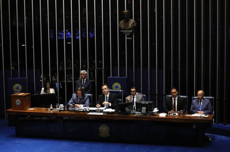 A imagem mostra uma sessão no Senado Federal do Brasil, com senadores sentados em mesas dispostas em formato de U. No fundo, há uma tela exibindo informações sobre a reunião. O ambiente é formal, com muitos participantes, incluindo senadores e assistentes, e a decoração é predominantemente azul.