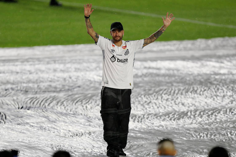 Fotografia mostra o jogador de futebol Neymar no estádio da Vila Belmiro. Ele está no gramado e aparece com os braços levantados， acenando para a torcida. Neymar usa calça preta， tênis preto e a camisa do Santos