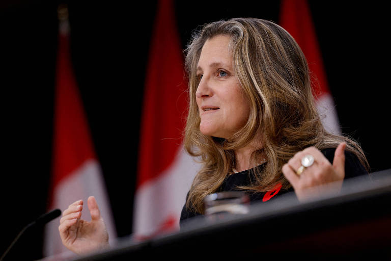 Uma mulher loira está falando em uma conferência de imprensa， com bandeiras do Canadá ao fundo. Ela usa uma blusa preta e gesticula com as mãos enquanto se expressa.
