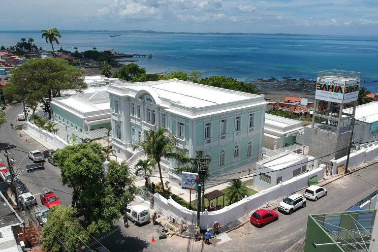 A imagem mostra um edifício de estilo arquitetônico clássico， pintado em tons de verde e branco， localizado em uma área costeira. O edifício possui janelas grandes e uma escadaria na entrada. Ao fundo， há uma vista do mar e do céu com algumas nuvens. Na parte inferior da imagem， é possível ver uma rua com carros estacionados e pessoas trabalhando em obras. À direita， há um letreiro que indica 039;Governo do Estado da Bahia039;.