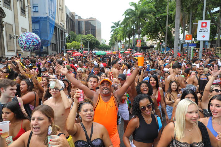 Uma grande multidão celebra o carnaval em uma rua， com pessoas vestindo roupas de verão e algumas sem camisa. Há um homem no centro， usando uma camiseta laranja e sorrindo， levantando um copo. Ao fundo， é possível ver árvores， prédios e barracas de comida. A atmosfera é festiva， com muitas pessoas dançando e se divertindo.