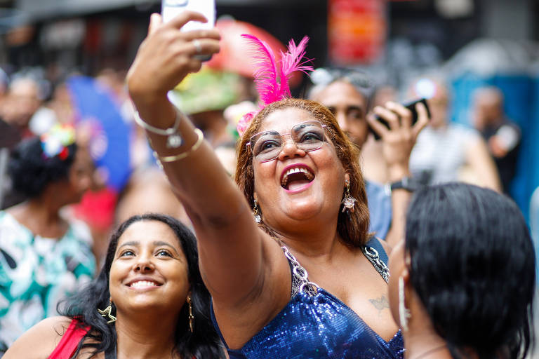 A imagem mostra um grupo de pessoas se divertindo durante uma festa de Carnaval. Uma mulher em destaque, com cabelo cacheado e uma fantasia brilhante azul, está tirando uma selfie com um smartphone, sorrindo amplamente. Ela usa um adereço de penas rosa na cabeça. Ao seu lado, outra mulher sorri para a câmera. O fundo é composto por outras pessoas e elementos festivos.