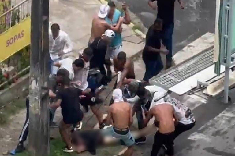 A imagem mostra um grupo de pessoas em uma rua， algumas delas envolvidas em uma briga ou confronto. Há várias pessoas em pé e outras no chão， com algumas sem camisa. Um homem está caminhando ao fundo， enquanto os demais parecem estar em uma situação de tensão. O ambiente urbano é visível， com postes de luz e fios elétricos ao fundo.