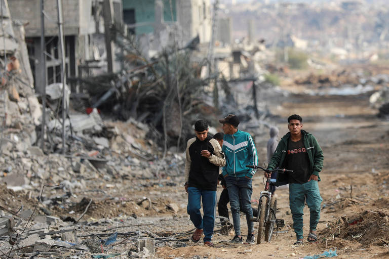 Três homens jovens caminham， um deles levando uma bicicleta. Ao fundo， desfocado， veem-se prédios destruídos e destroços