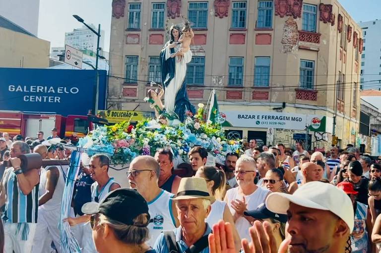 A imagem mostra uma multidão reunida em uma rua durante uma festa religiosa. No centro， há uma grande imagem de uma figura religiosa， cercada por flores e decorada com elementos festivos. As pessoas estão vestidas de maneira variada， algumas usando chapéus e outras com roupas coloridas. Ao fundo， é possível ver prédios e uma loja com o nome 039;Gallepalo Center039;. O céu está claro e ensolarado.