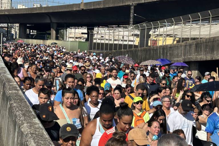 A imagem mostra uma multidão reunida em uma rua durante uma festa religiosa. No centro， há uma grande imagem de uma figura religiosa， cercada por flores e decorada com elementos festivos. As pessoas estão vestidas de maneira variada， algumas usando chapéus e outras com roupas coloridas. Ao fundo， é possível ver prédios e uma loja com o nome 039;Gallepalo Center039;. O céu está claro e ensolarado.