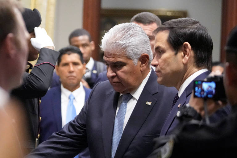 Homem idoso branco， de cabelos brancos， usando terno escuro， camisa branca e gravata cinza， aparece ao lado de homem de meia idade branco， de cabelos castanhos， terno escuro， os dois estão de perfil e há outras pessoas ao fundo