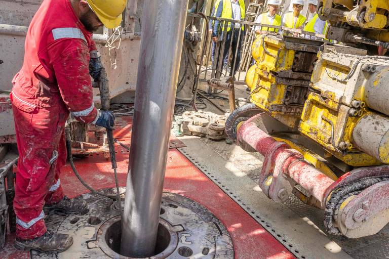 Um trabalhador vestido com um macacão vermelho e um capacete amarelo está operando uma máquina de perfuração. A máquina possui um tubo metálico vertical e está posicionada sobre uma base de concreto. Ao fundo， há outros trabalhadores e equipamentos de construção.