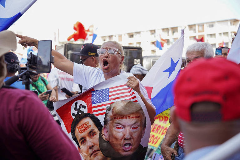 Mulher idosa branca de óculos e camiseta branca gesticula em meio a multidão com bandeiras; ela segura cartaz com bandeira dos EUA， símbolo nazista e fotos de Donald Trump e Marco Rubio