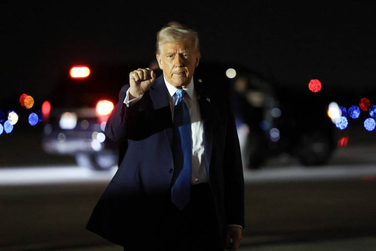 Um homem em traje formal， com cabelo loiro， está posando em um ambiente noturno. Ele levanta uma mão com o punho fechado. Ao fundo， há veículos com luzes acesas， sugerindo uma cena de segurança ou vigilância.
