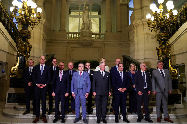 Um grupo de treze homens e mulheres posando em um ambiente interno elegante, com escadarias e iluminação decorativa. Eles estão vestidos formalmente, com ternos e gravatas, e estão organizados em duas fileiras. Ao fundo, há uma escultura ou estátua visível, além de grandes luminárias.