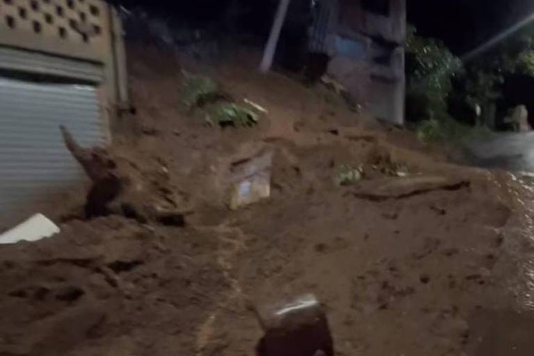 A imagem mostra um deslizamento de terra em uma área urbana durante a noite. O solo está coberto de lama e detritos， com alguns objetos， como caixas， visíveis. Ao fundo， há uma construção com uma parede e uma porta， e a iluminação da rua é fraca， sugerindo que é de noite.