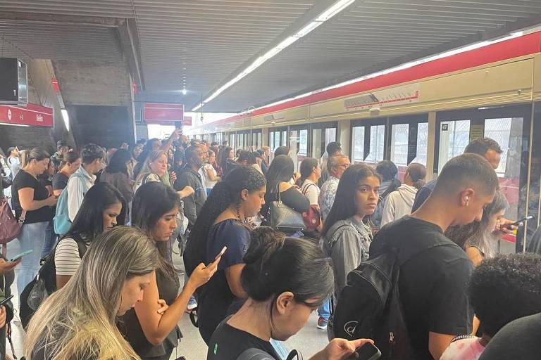 A imagem mostra uma estação de metrô lotada， com um grande número de pessoas aguardando na plataforma. A maioria dos indivíduos está olhando para seus celulares. O ambiente é iluminado， com luz natural entrando pela parte superior. Ao fundo， pode-se ver o trem do metrô， que está parado na plataforma.