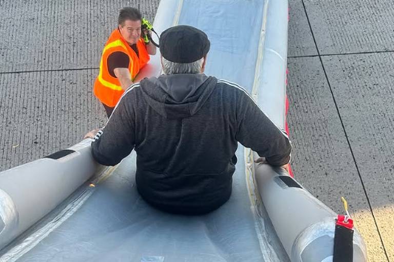 A imagem mostra um homem sentado em um bote inflável， que está posicionado em uma superfície de concreto. Ele está usando um cap e uma roupa escura. Dois homens estão próximos a ele: um está vestindo um colete laranja e o outro é um bombeiro com uniforme amarelo. O ambiente parece ser uma área de desembarque de um aeroporto.