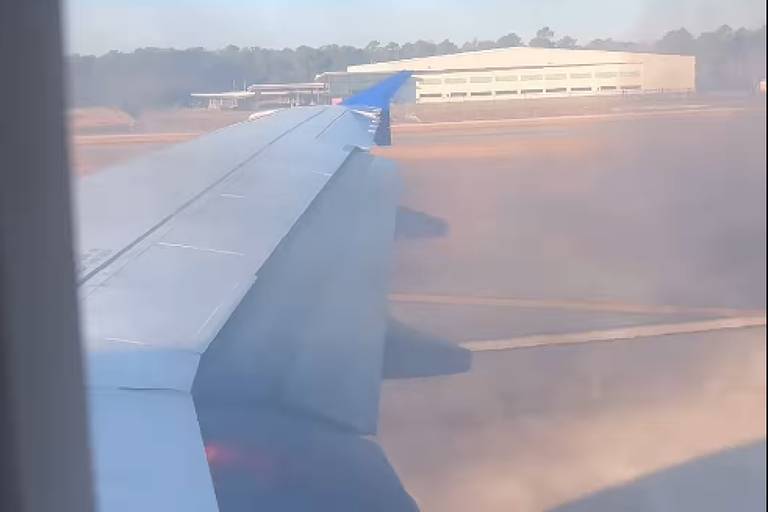 A imagem mostra a asa de um avião vista pela janela， com um fundo de um aeroporto. O céu está claro e há uma pista de pouso visível ao fundo， além de um edifício de hangar.