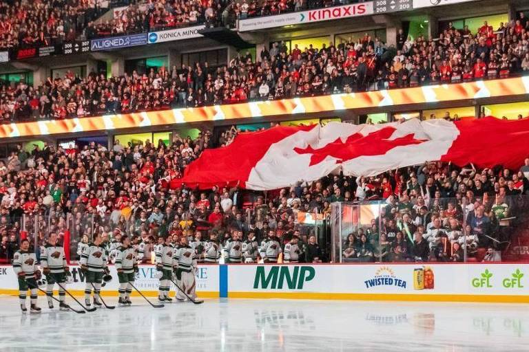 A imagem mostra uma grande bandeira do Canadá sendo exibida por torcedores em um estádio de hóquei. A multidão está vestida em roupas vermelhas e brancas， apoiando a equipe. No gelo， jogadores estão alinhados， possivelmente durante a execução do hino nacional. O ambiente é vibrante e cheio de energia.