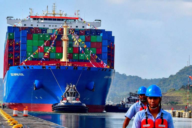 A imagem mostra um grande navio de carga colorido， com contêineres em várias cores， se aproximando de um porto. Na frente do navio， há um rebocador e， em primeiro plano， dois homens usando uniformes azuis e capacetes， caminhando em direção ao navio. O fundo apresenta uma paisagem montanhosa e céu claro.