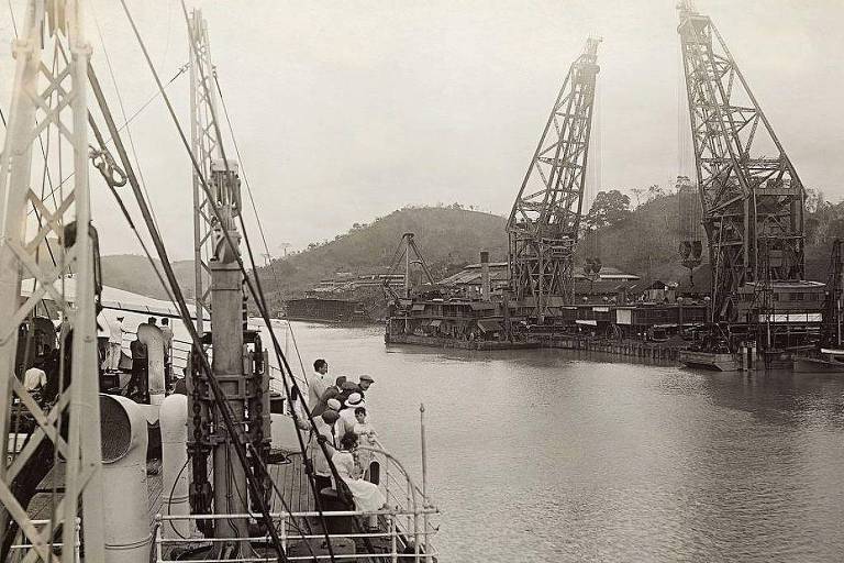 A imagem mostra um porto com guindastes altos e estruturas de madeira ao fundo. No primeiro plano， há um barco com várias pessoas em sua borda， observando a cena. O ambiente é montanhoso， com colinas ao fundo e um céu nublado.