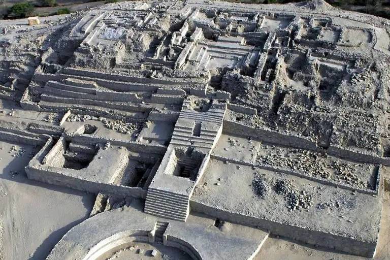 Imagem aérea de uma antiga cidade em ruínas， com estruturas de pedra visíveis， incluindo edifícios， praças e caminhos. O terreno ao redor é árido e a vegetação é escassa.