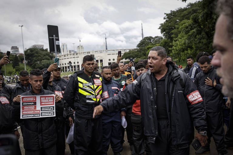 A imagem mostra um grupo de homens， alguns em uniformes de segurança， em uma manifestação. Um dos homens， que parece ser um líder， está gesticulando enquanto fala. Outros seguram cartazes. Ao fundo， há um edifício e árvores， com um céu nublado.