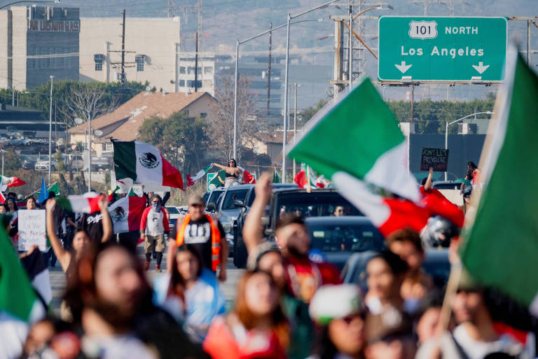 A imagem mostra uma multidão de pessoas em um protesto， segurando bandeiras do México. Ao fundo， há um sinal de trânsito indicando a direção para Los Angeles， com a rodovia 101 Norte. Vários carros estão parados na estrada， e a atmosfera parece ser de celebração ou manifestação.