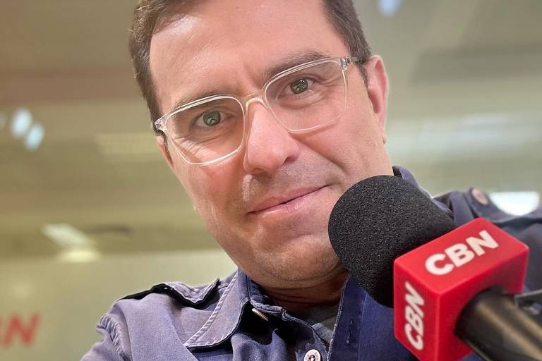 Um homem está posando para a câmera em um estúdio de rádio. Ele usa óculos e uma camisa azul escura. O microfone próximo a ele tem um logotipo vermelho com a inscrição 'CBN'. Ao fundo, é possível ver uma iluminação suave e parte do estúdio.