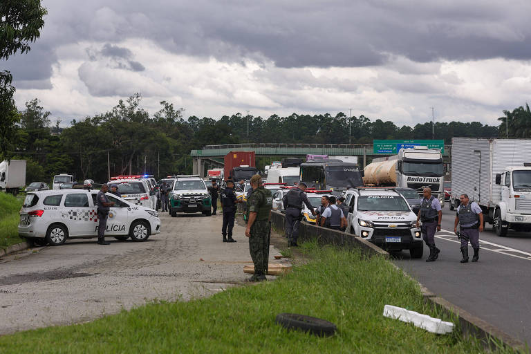 A imagem mostra uma operação policial em uma estrada， com várias viaturas da polícia e caminhões ao fundo. Há um grupo de pessoas， incluindo policiais e civis， em atividade na cena. O céu está nublado， e a vegetação é visível ao lado da estrada.