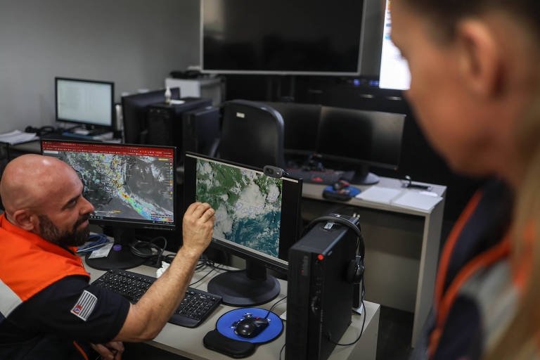 A imagem mostra um homem com uma camisa de trabalho laranja e preta， sentado em frente a um computador， apontando para uma tela que exibe dados meteorológicos. Ao fundo， há outros monitores e uma mulher com cabelo preso， observando a tela. O ambiente é um centro de monitoramento.