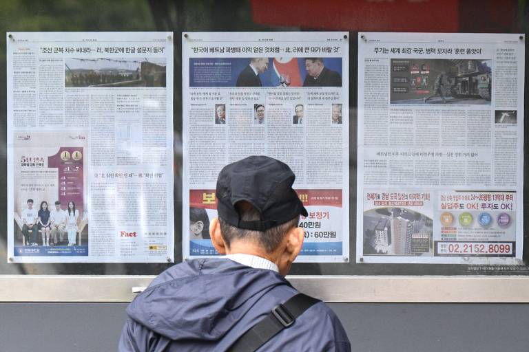 Um homem de costas， usando um boné e uma jaqueta escura， está parado em frente a um painel com várias páginas de jornais expostas. As páginas contêm textos e imagens， mas não é possível ler o conteúdo. O ambiente parece ser urbano， com um foco na leitura de notícias.
