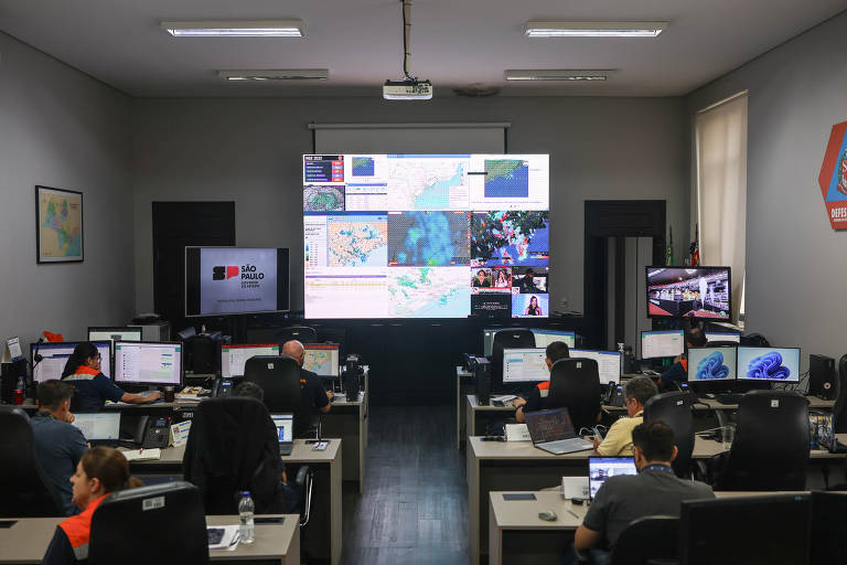 A imagem mostra uma sala de controle com várias mesas e computadores. No fundo, há uma tela grande exibindo mapas e dados. Várias pessoas estão sentadas em suas mesas, olhando para os monitores. A sala é bem iluminada e possui paredes claras.