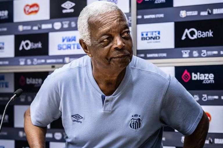 Um homem de pele escura， com cabelo grisalho， está sentado em uma mesa de conferência. Ele usa uma camisa azul clara com o emblema de um time de futebol. Na mesa， ele segura um par de luvas de goleiro. Ao fundo， há um painel com logotipos de patrocinadores.