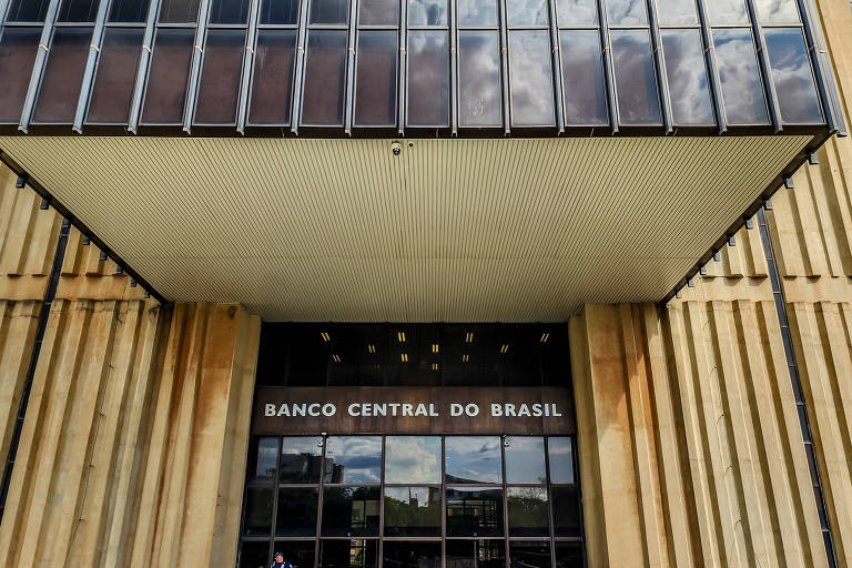 A imagem mostra a fachada do Banco Central do Brasil， com uma entrada principal visível. O edifício possui uma estrutura de madeira e vidro， com um toldo projetado na parte superior. O nome 039;BANCO CENTRAL DO BRASIL039; está claramente visível na entrada.
