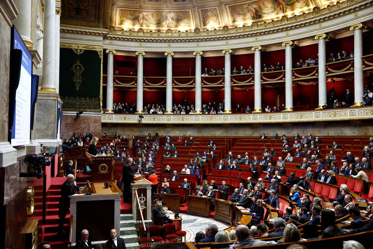 A imagem mostra o interior de um grande salão legislativo， com várias fileiras de assentos ocupados por pessoas. O ambiente é decorado com colunas brancas e detalhes em vermelho， e há um palco à frente onde um orador está se apresentando. O público parece atento， e há uma tela ao fundo.