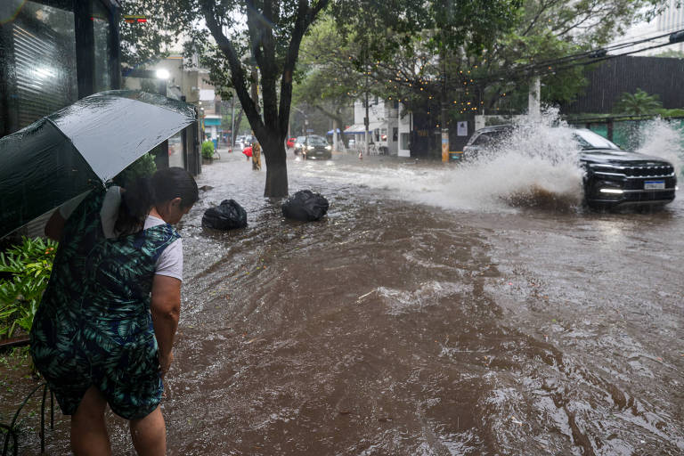 A imagem mostra uma cena de inundação em uma área urbana. Uma mulher， segurando um guarda-chuva， está em primeiro plano， observando a água que cobre a calçada e parte da rua. Ao fundo， um carro passa， levantando água. Há árvores e sacos de lixo visíveis na cena， e o céu parece nublado， indicando chuva.