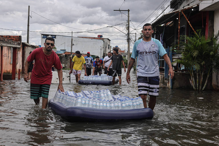 A imagem mostra várias pessoas caminhando em uma área inundada， com água até os joelhos. Algumas delas estão puxando botes infláveis com garrafas de água mineral. 