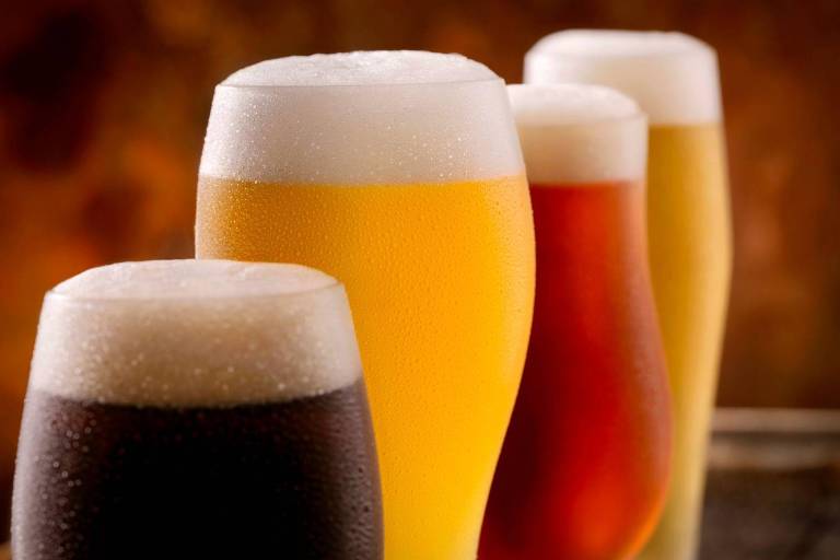 A imagem mostra quatro copos de cerveja em diferentes cores e estilos. O primeiro copo à esquerda contém uma cerveja escura， seguido por um copo com cerveja clara， um copo com cerveja âmbar e， por último， um copo com cerveja dourada. Todos os copos têm uma camada de espuma no topo.