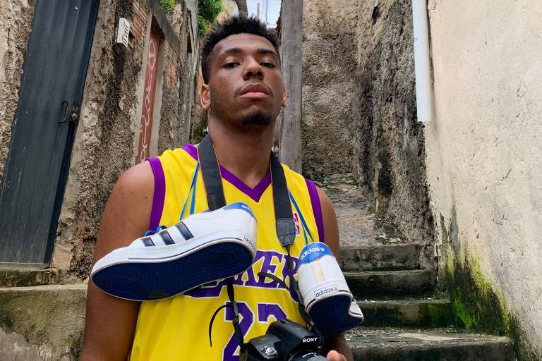 Um jovem está posando em uma escadaria de uma área urbana. Ele usa uma camiseta amarela com detalhes roxos e o número 27. Em seu pescoço， ele carrega um tênis branco com detalhes azuis e uma câmera fotográfica preta na mão. Ao fundo， há uma parede de pedra e uma árvore com folhas verdes. O céu está nublado.