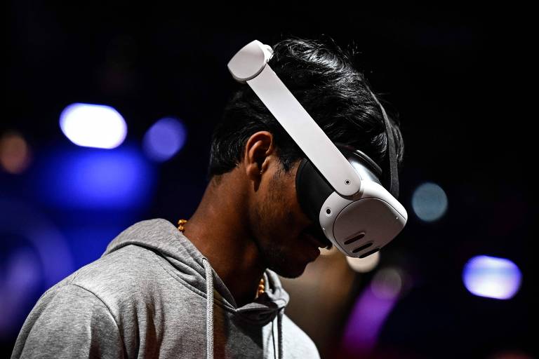 A imagem mostra uma pessoa usando um headset de realidade virtual. A pessoa está de perfil， com a cabeça ligeiramente inclinada para baixo. O ambiente ao fundo é desfocado， com luzes coloridas que sugerem um evento ou uma feira de tecnologia.
