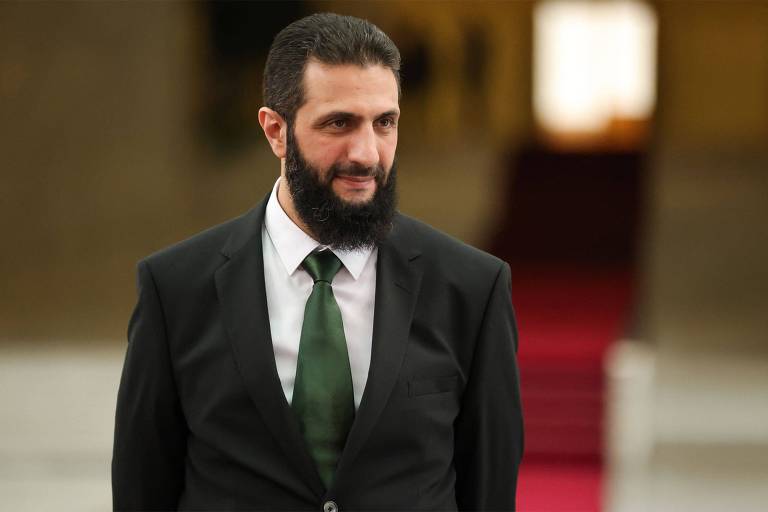 Um homem com barba e cabelo escuro， vestindo um terno preto e uma gravata verde， está em um ambiente interno com um fundo desfocado. O chão parece ser de mármore e há um tapete vermelho visível ao fundo.