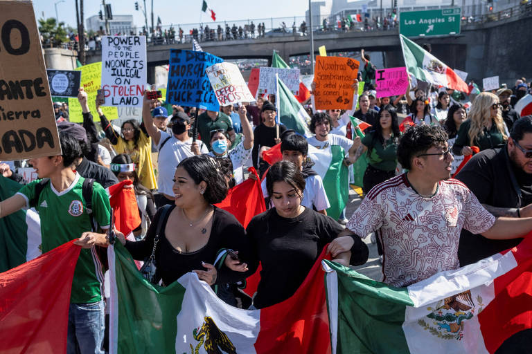 A imagem mostra um grupo de pessoas participando de um protesto. Elas seguram bandeiras do México e cartazes coloridos. O ambiente é ao ar livre， com uma estrada ao fundo e bandeiras mexicanas visíveis. Algumas pessoas estão usando máscaras. O clima parece ser de mobilização e união