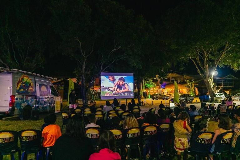 A imagem mostra uma exibição de filme ao ar livre durante a noite. Um projetor exibe uma cena de um filme em uma tela grande, enquanto várias cadeiras estão dispostas em frente a ela, ocupadas por um público diversificado. À esquerda, há uma van decorada com cores vibrantes e o nome 'CINESOLAR'. Ao fundo, é possível ver árvores, iluminação de um estabelecimento comercial e veículos estacionados.