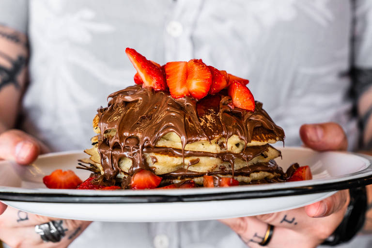 Uma pessoa está segurando um prato com uma pilha de panquecas cobertas com chocolate derretido e morangos fatiados no topo. O fundo é desfocado, mas é possível ver uma camisa clara da pessoa que segura o prato.