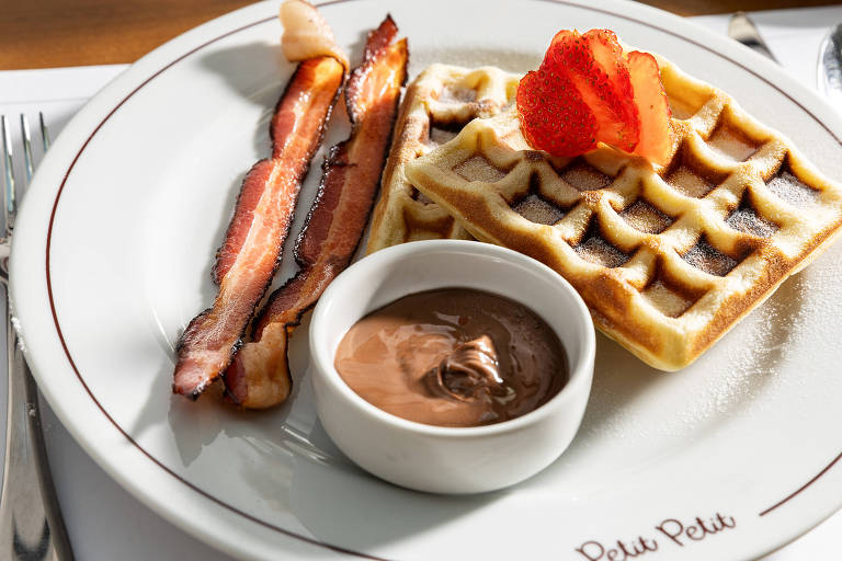 Um prato de café da manhã apresenta waffles, tiras de bacon crocante, um pequeno recipiente com creme de chocolate e morangos fatiados. O prato é decorado com uma borda vermelha e a inscrição 'Petit Petit'.